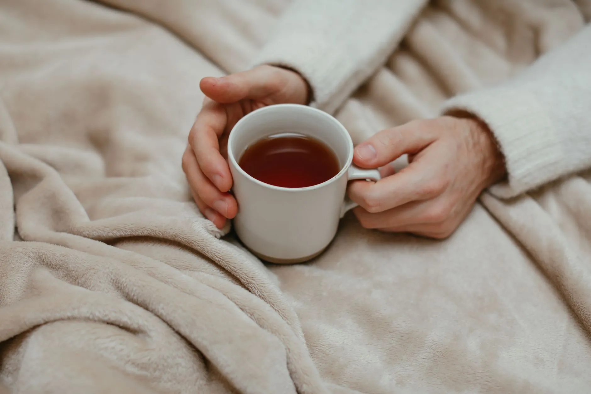 Hands holding a mug of tea over a soft beige blanket.