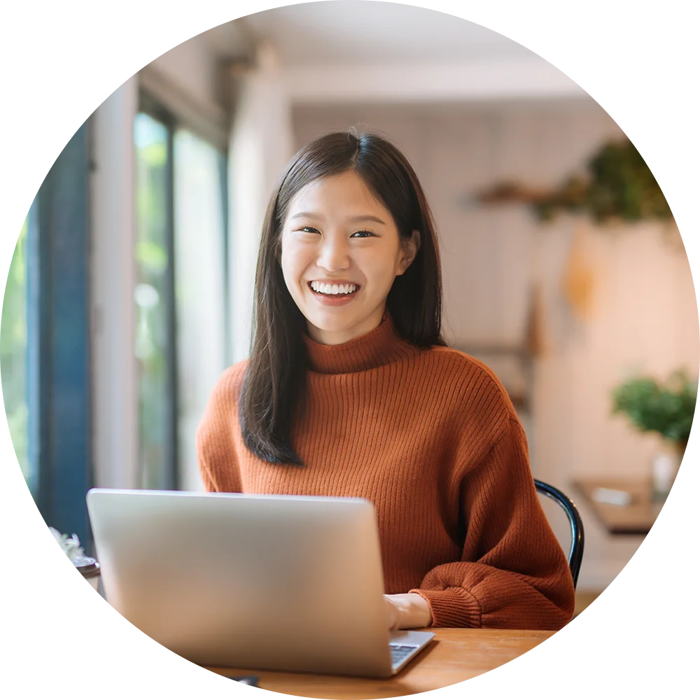 A smiling young woman uses a laptop at a table. She enjoys work and life after addiction treatment at Jasper Grove Recovery.
